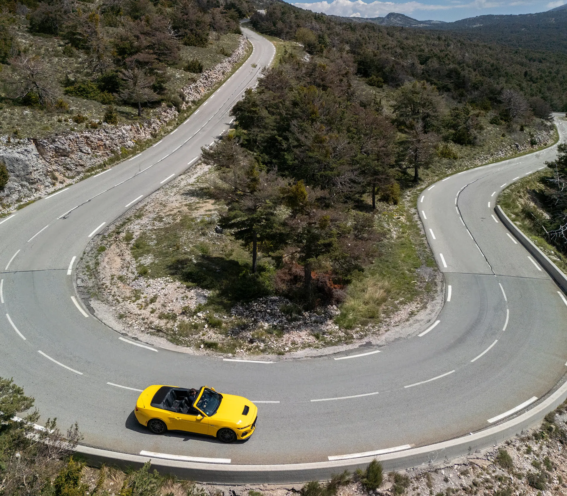 gelber Mustang als Cabrio fährt auf einer kurvigen Straße aus der Vogelperspektive