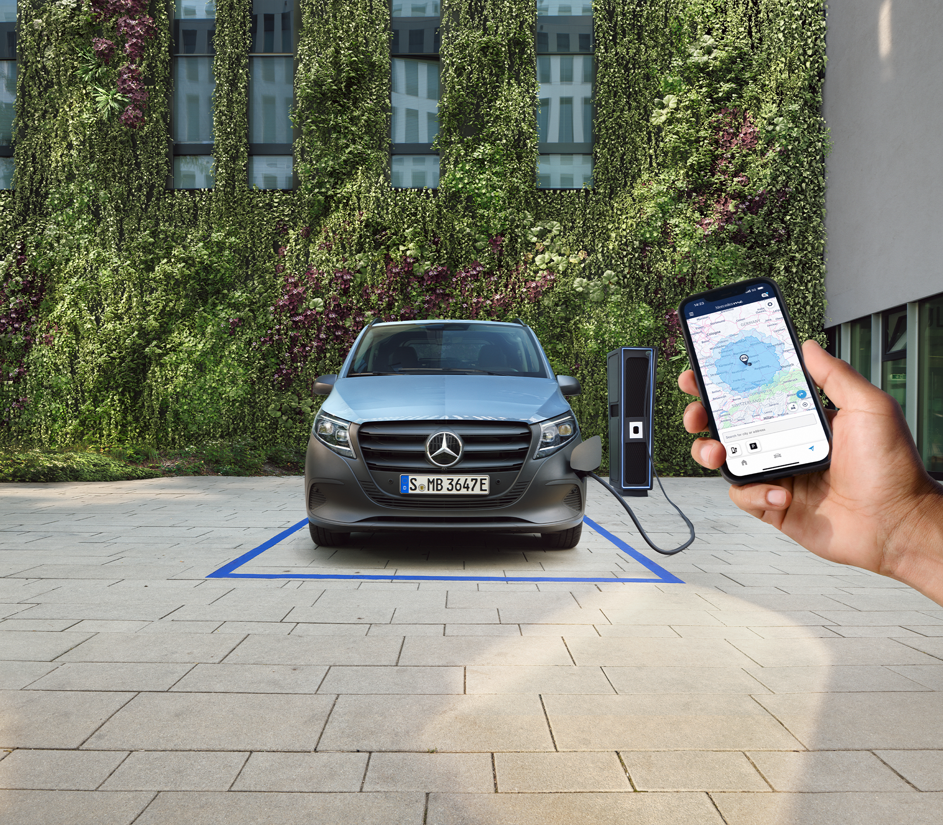 Vorderansicht eines Mercedes-Benz eVito beim Ladevorgang vor einer begrünten Fassade. Im Vordergrund hält eine Hand ein Smartphone mit Navigations-App.