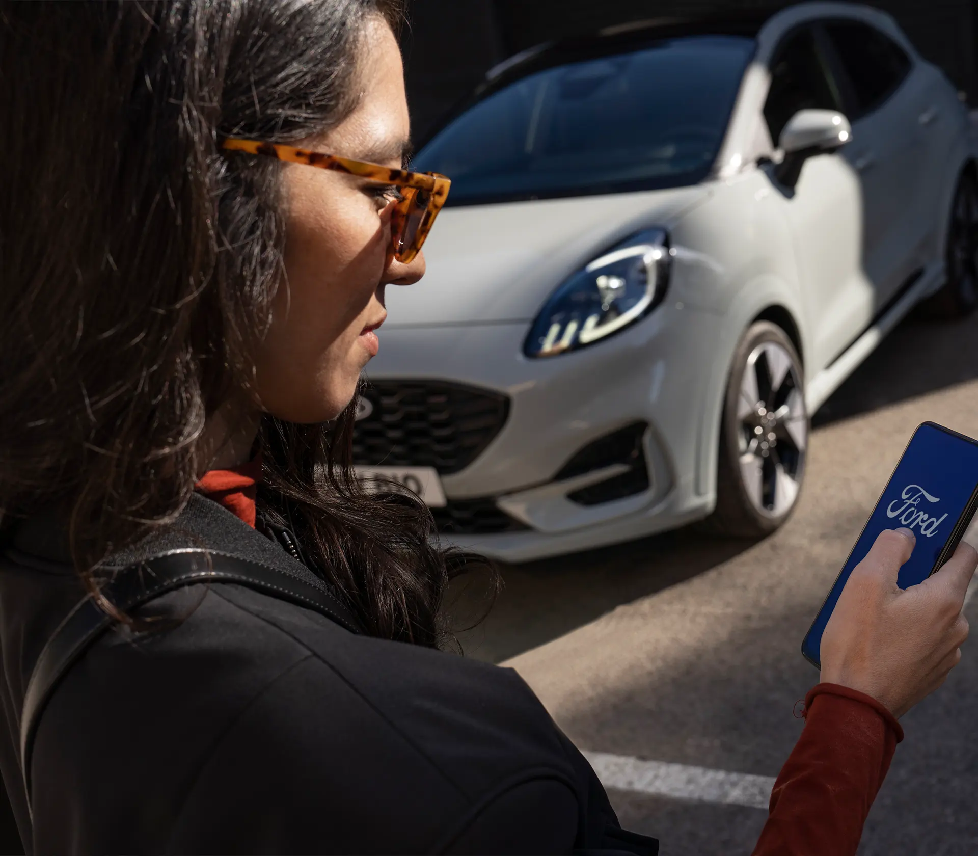 Frau bedient Ihr Handy mit dem Ford Logo drauf, im Hintergrund steht der graue Puma geparkt