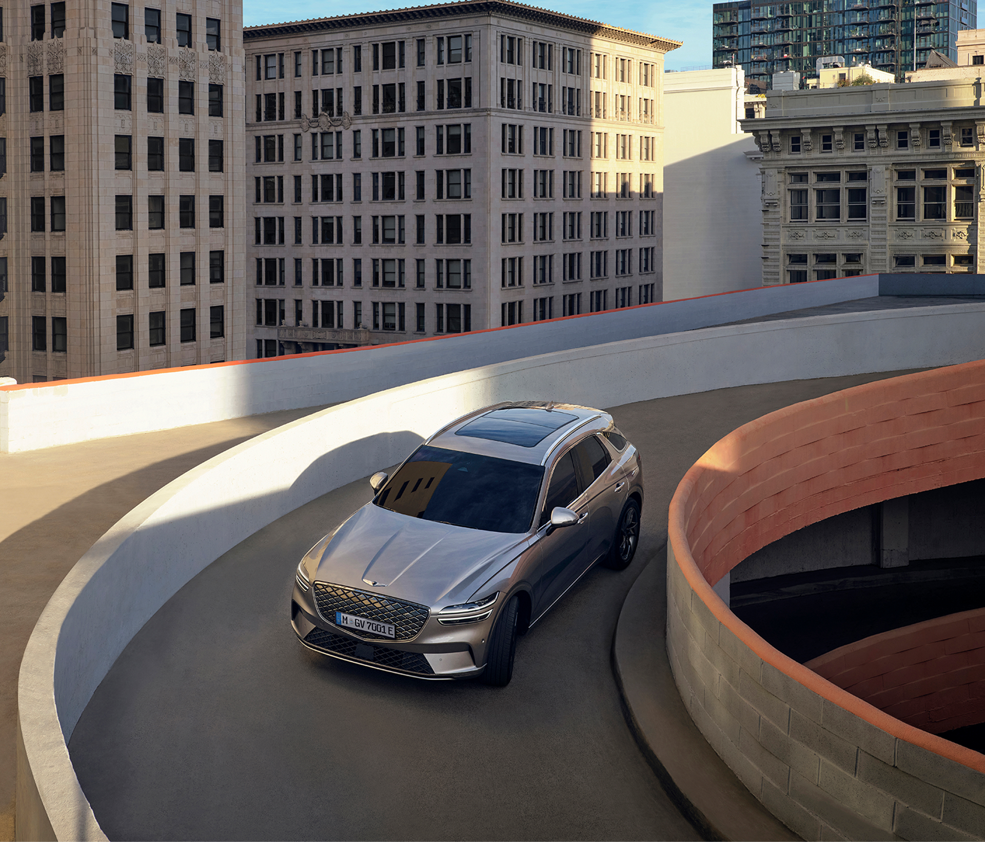 Genesis Electrified GV 70 in grau fährt von einem Parkdeck in einer engen kurvigen Straße runter. Im Hintergrund sind Gebäude.