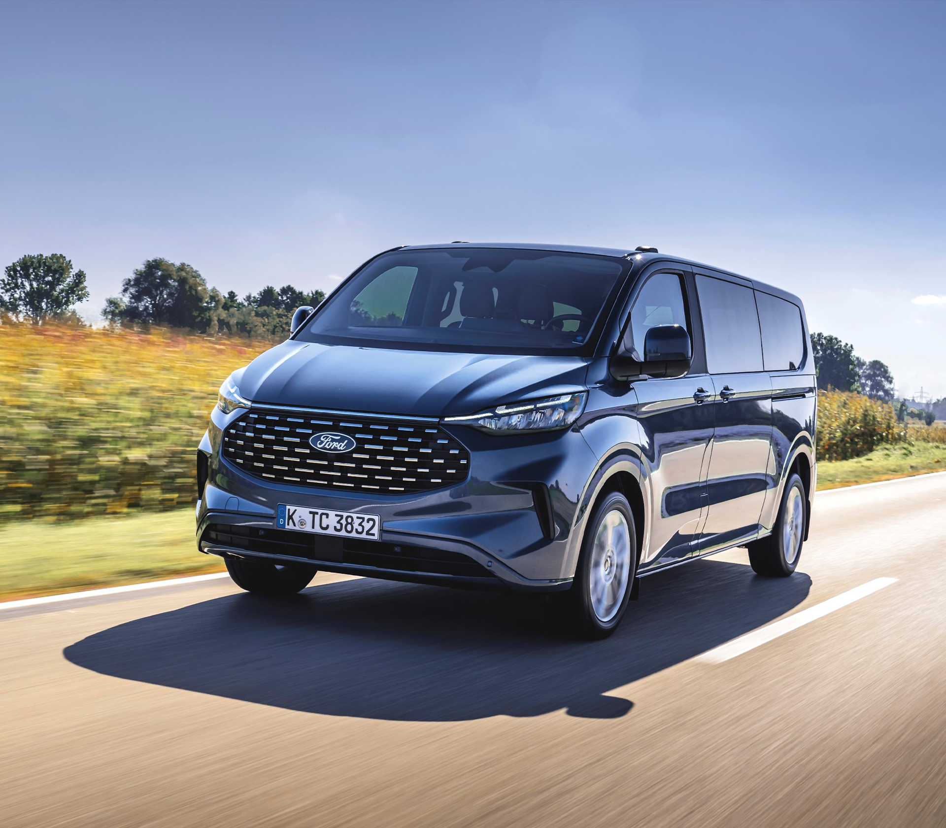 Ford Tourneo Custom Frontanischt in lau. Auto fährt auf einer Straße mit grünenfeldern und Bäumen daneben und im Hintergrund.