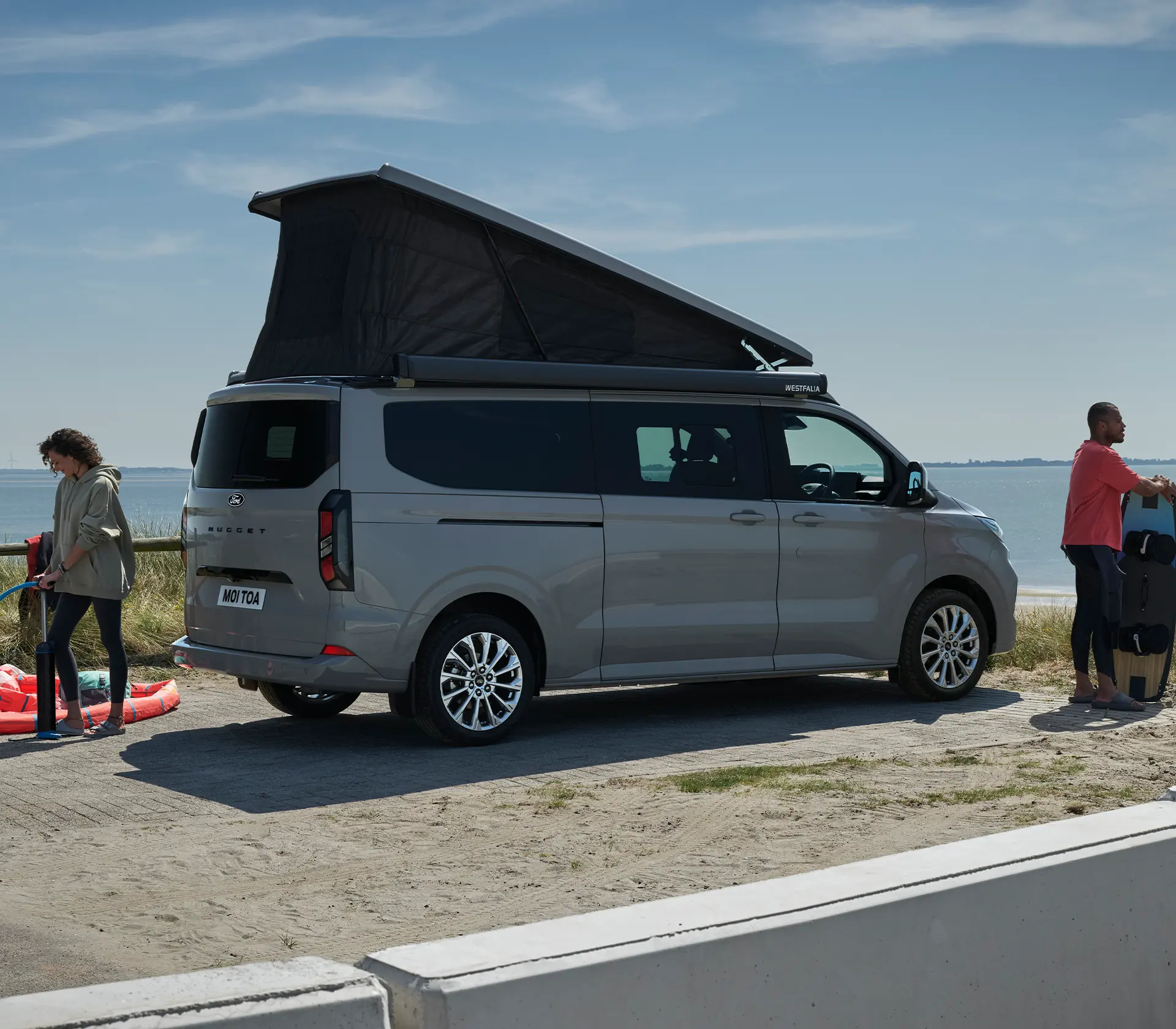 Grauer Ford Nugget mit offenem Dach - Mann und Frau daneben bereite sich aufs Surfen vor
