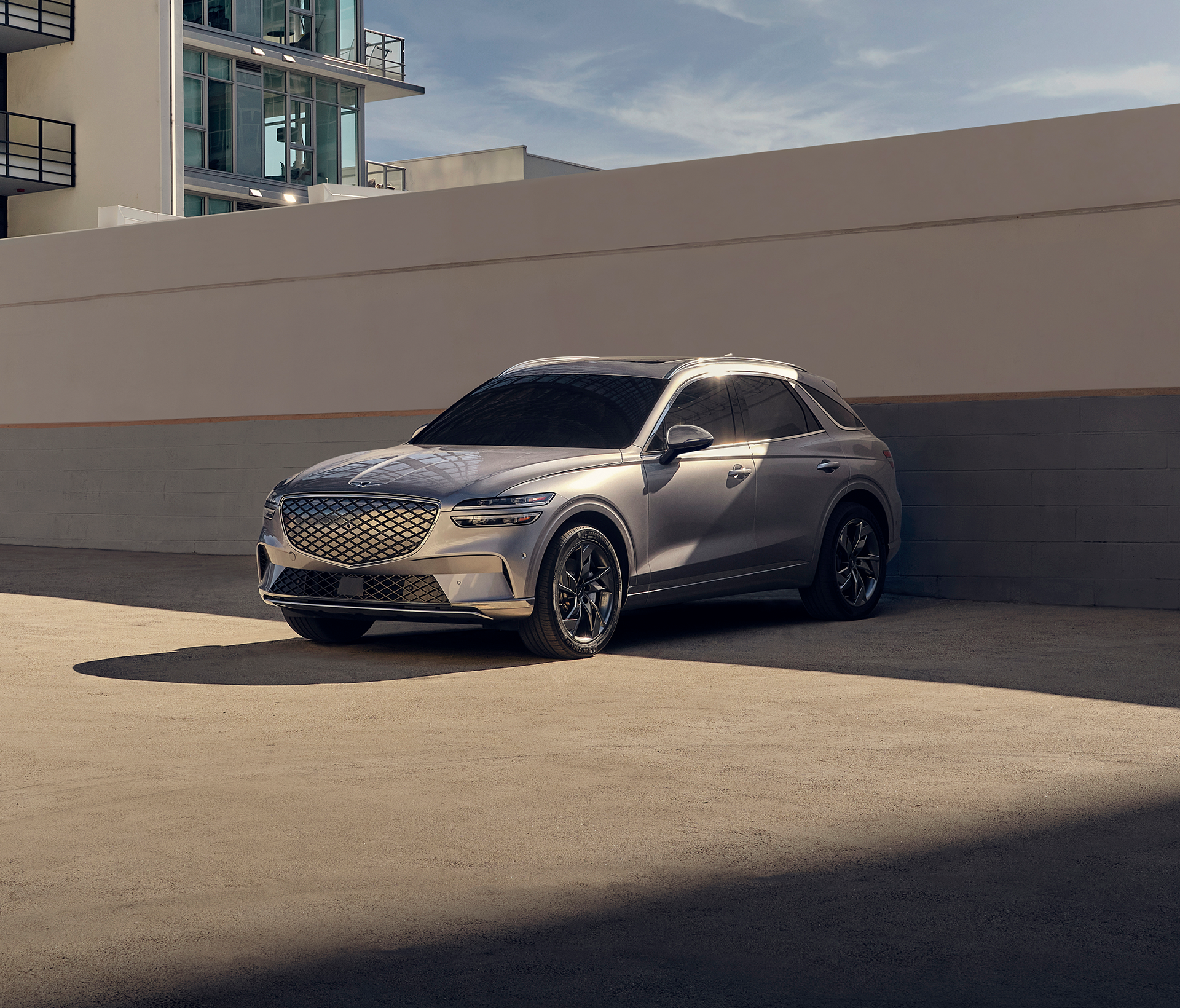 Genesis Electrified GV 70 in grau steht vor einer weiß/grauen Wand geparkt mit dem Heck zur Wand gerichtet. Über der Wand erkennt man etwas blauen Himmel und ein modernes Gebäude am Rand des Bildes.