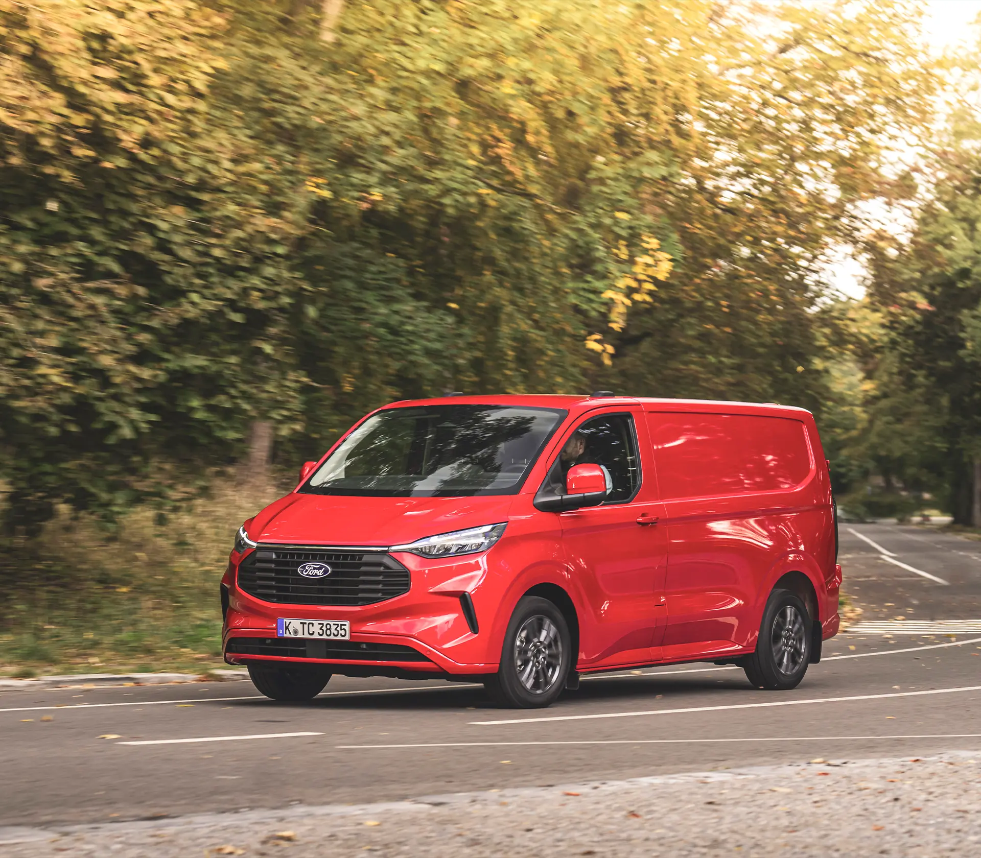 seitliche Ansicht auf roten transit-Custom welcher auf einer Straße umgeben von vielen Bäumen fährt