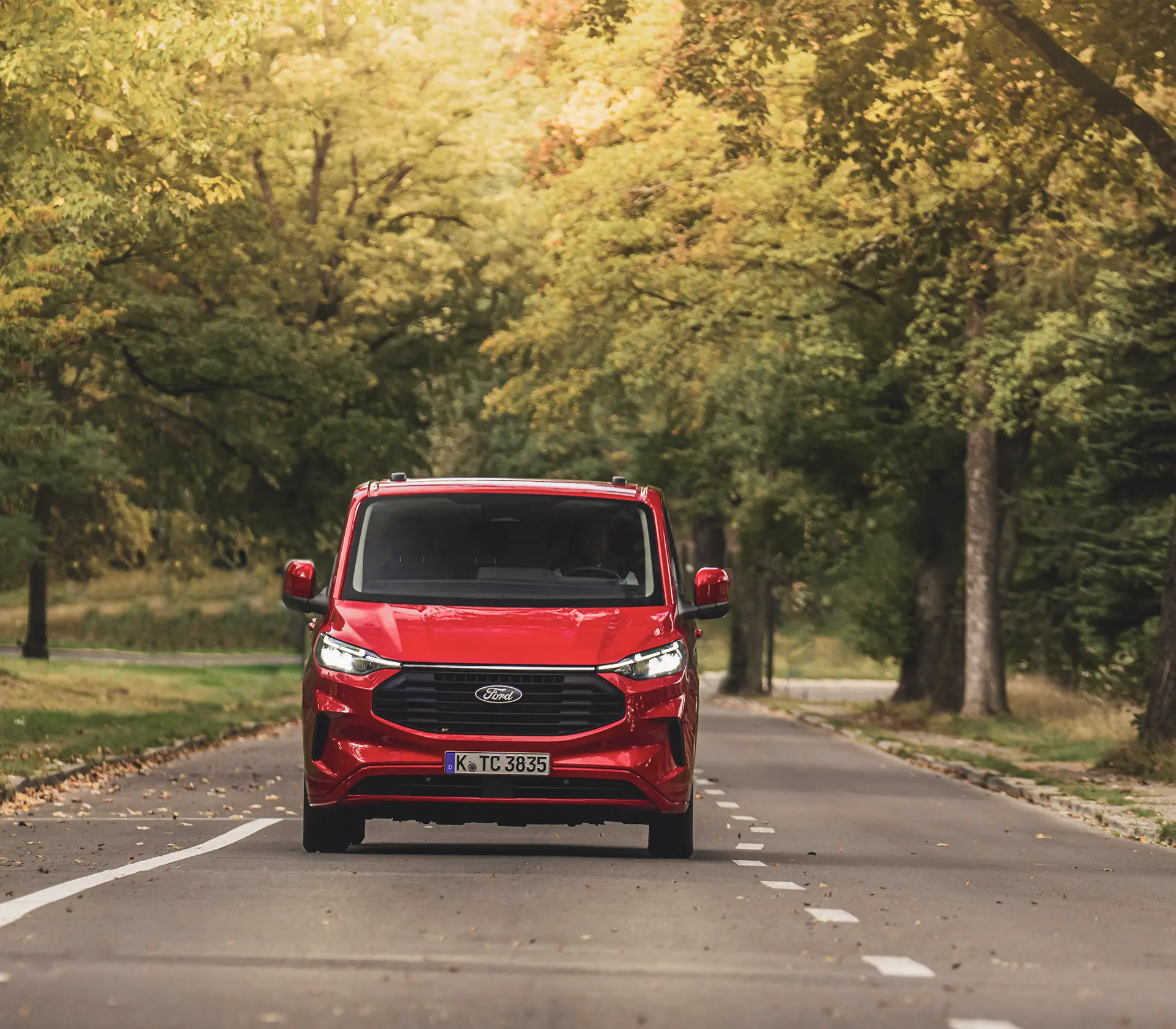 Frontansicht auf roten transit-Custom welcher auf einer Straße umgeben von vielen Bäumen fährt