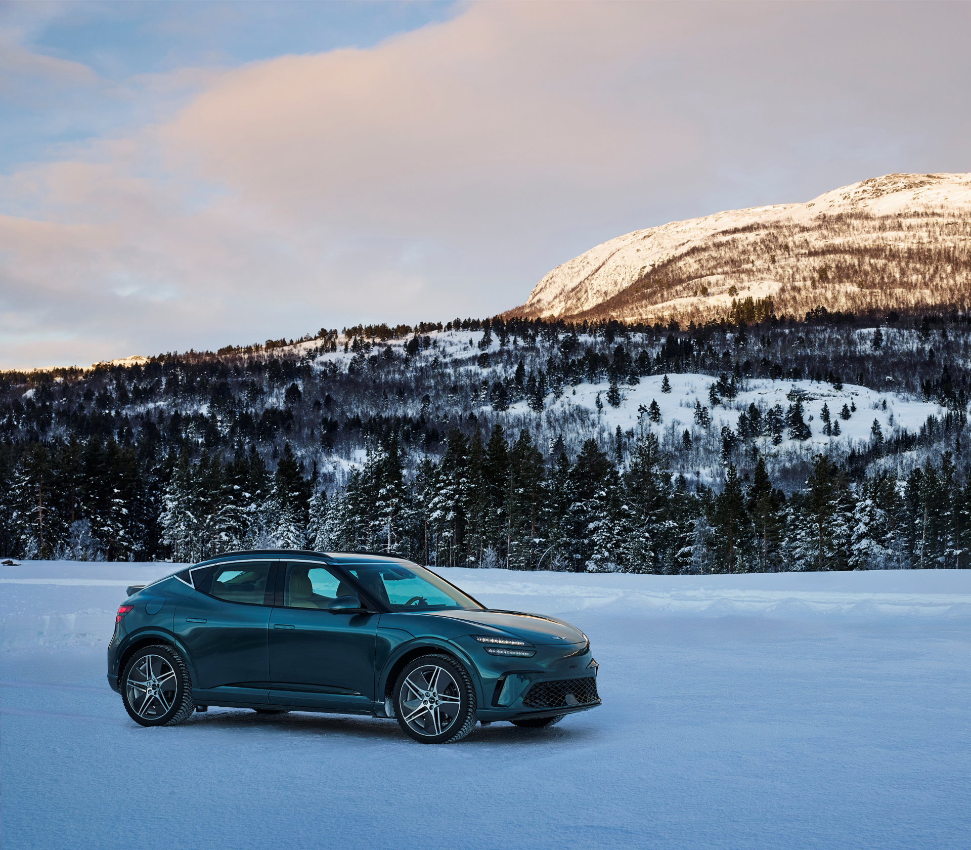 Genesis GV 60 Seitenansicht im Schnee mit schneebedecktem Wald und Bergen im Hintergrund