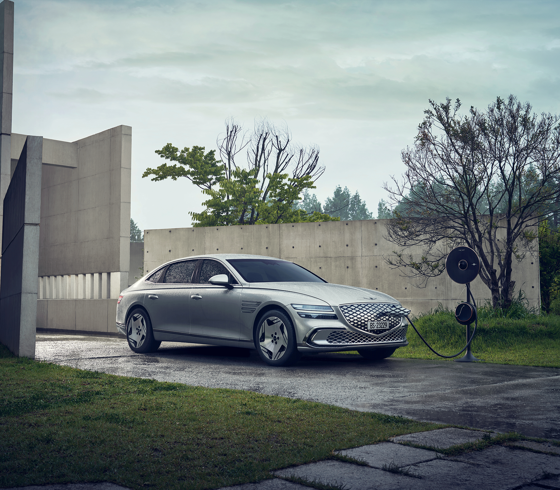 Genesis Electrified G 80 steht in einer verregneten gegend mit einer Mauer und einigen Bäumen während das graue Auto lädt