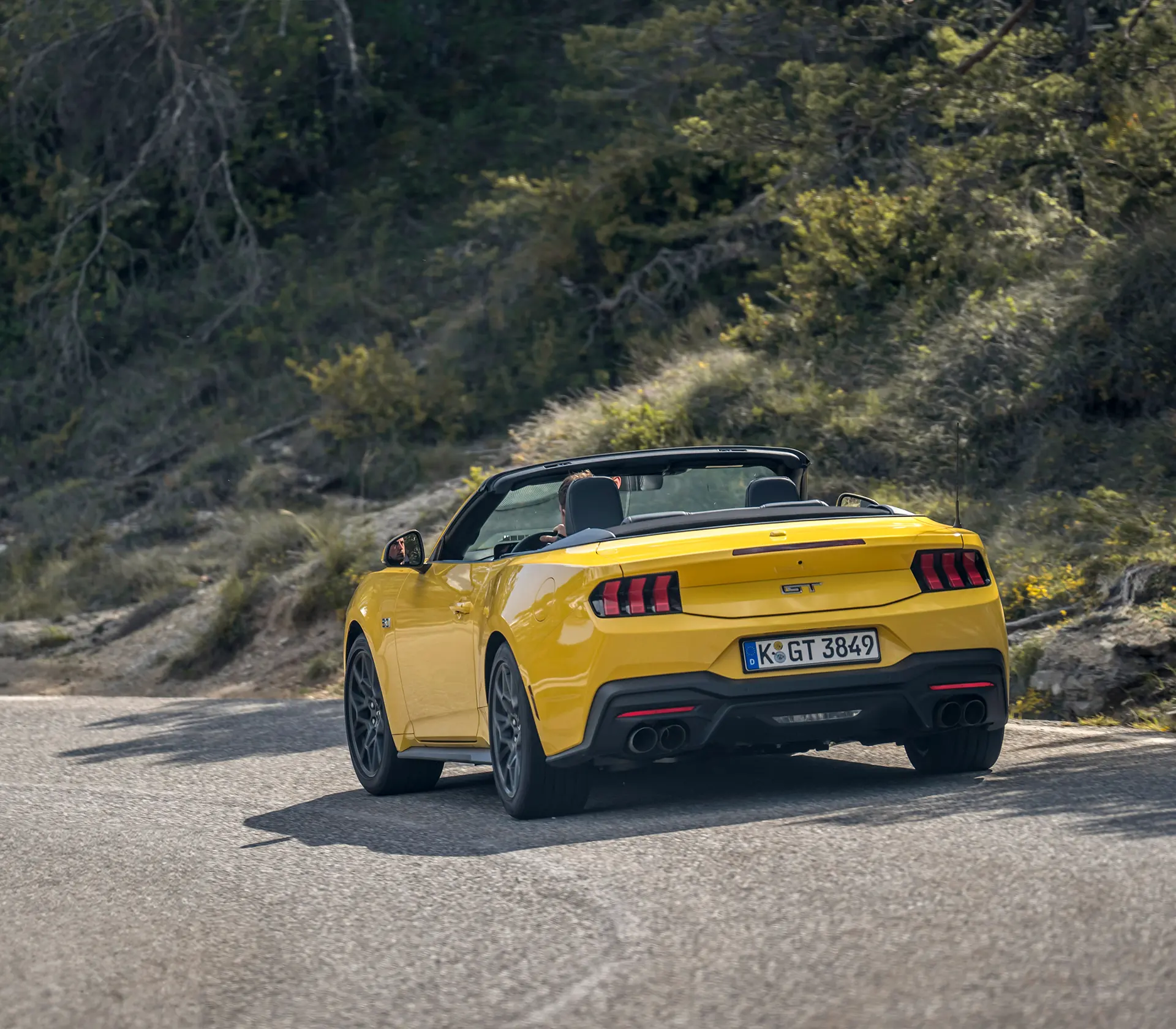 Heckansicht auf gelber Mustang als Cabrio fährt auf einer kurvigen Straße