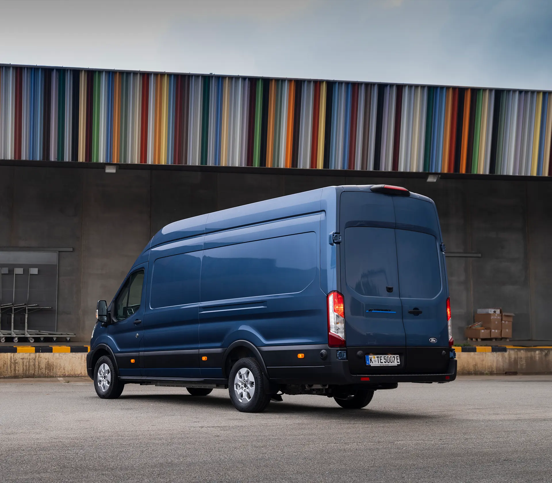 seitliche Heckansicht auf einen blauen E-Transit vor einem buntem Dach