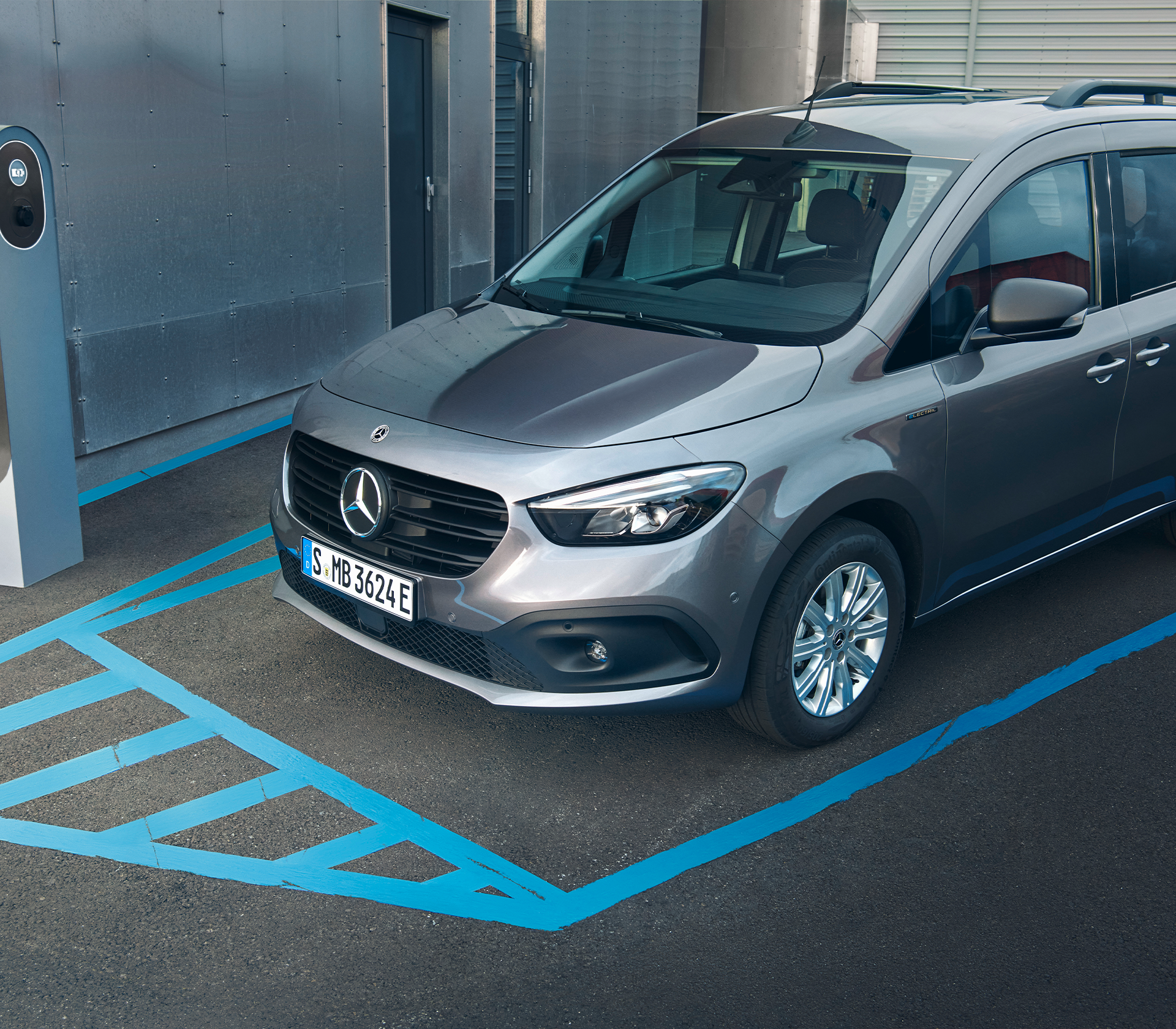 Grauer Mercedes-Benz eCitan an einer Ladestation, geparkt auf einem blau markierten E-Mobilitätsparkplatz neben Industriegebäude.