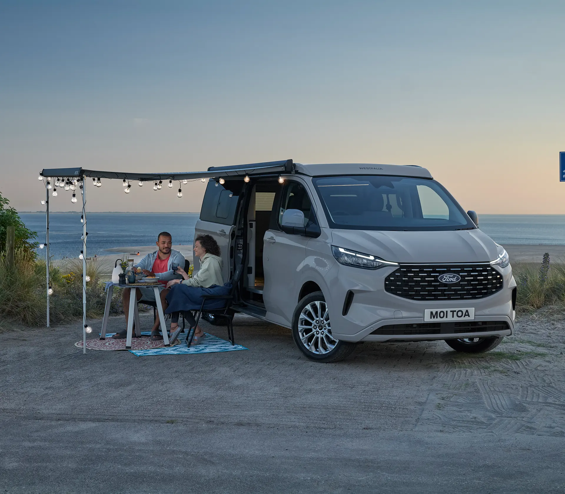 Mann und Frau sitzen vor dem Ford Nugget unter einer Plane und Essen gemeinsam im Sonnenuntergang