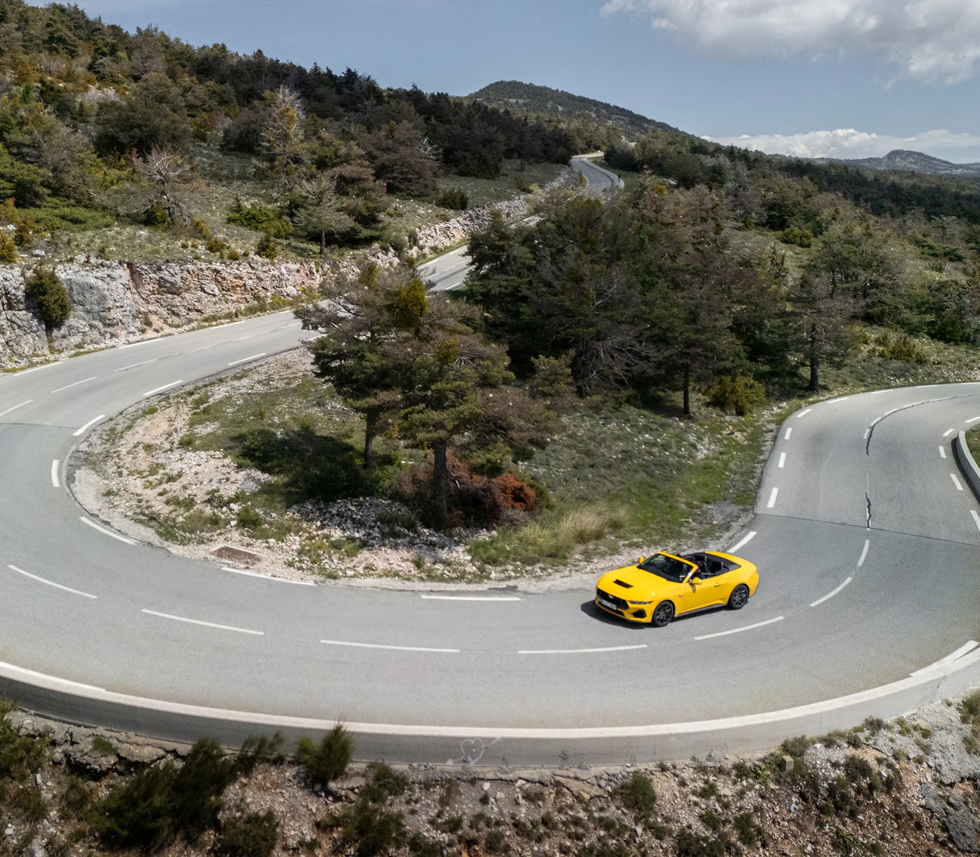 gelber Mustang als Cabrio fährt auf einer kurvigen Straße aus der Vogelperspektive