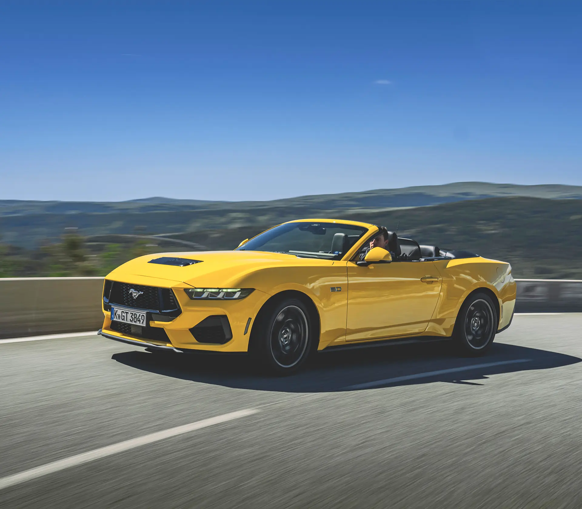 gelber Mustang als Cabrio fährt auf einer kurvigen Straße