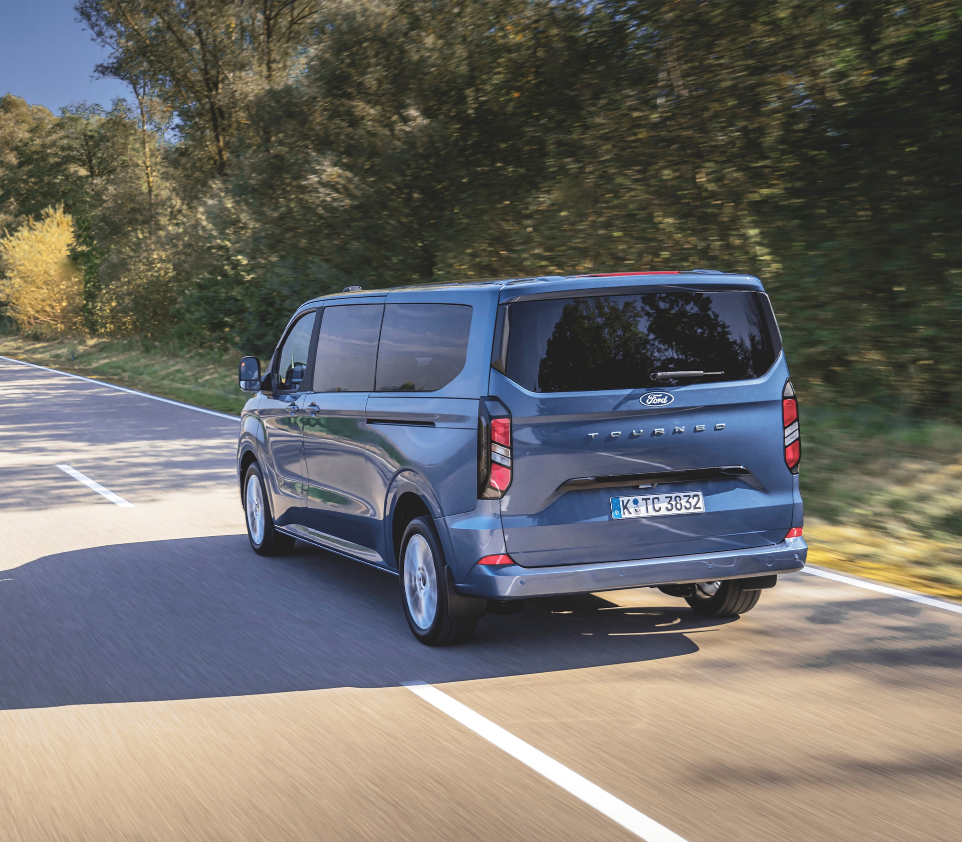 Ford Tourneo Custom Heckansicht in Blau fährt auf einer Straße mit Bäumen im Hintergrund