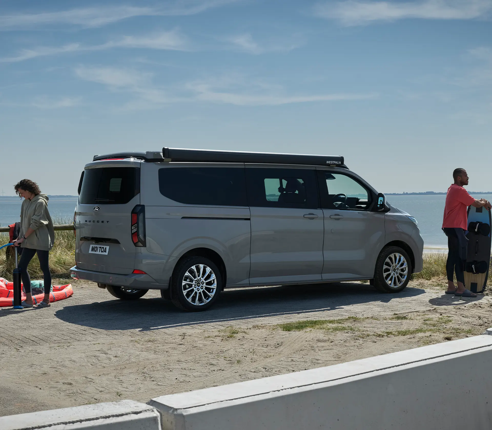Grauer Ford Nugget mit geschlossenem Dach - Mann und Frau daneben bereite sich aufs Surfen vor