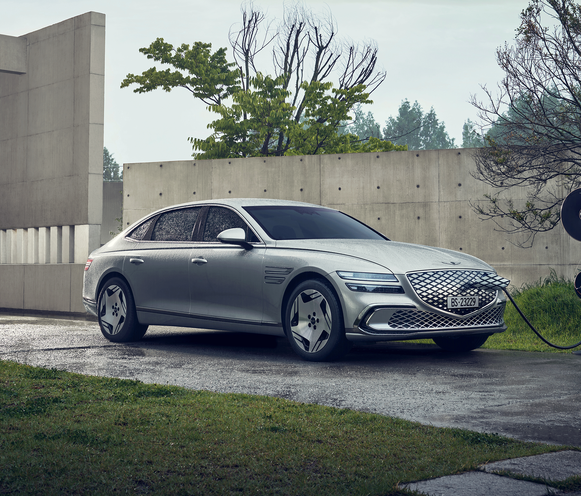 Genesis Electrified G 80 steht in einer verregneten gegend mit einer Mauer und einigen Bäumen während das graue Auto lädt